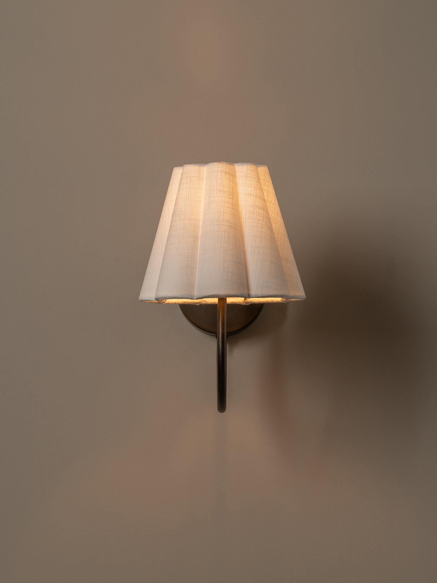 A wall-mounted sconce with a pleated white fabric shade and a dark metal arm, casting a warm light against a plain taupe wall.