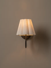 A wall-mounted sconce with a pleated white fabric shade and a dark metal arm, casting a warm light against a plain taupe wall.