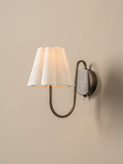 A wall-mounted sconce with a pleated white fabric shade and a curved dark metal arm, shown lit against a plain beige wall.