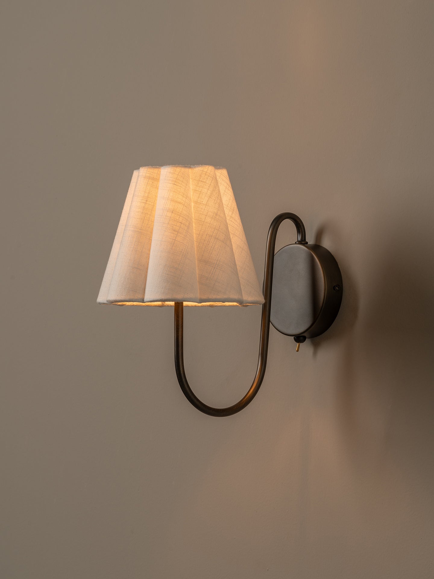 A modern wall sconce with a pleated white fabric shade and a curved dark metal arm, mounted on a warm beige wall and illuminated.