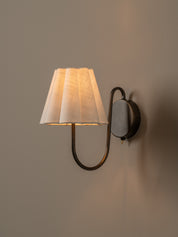 A modern wall sconce with a pleated white fabric shade and a curved dark metal arm, mounted on a warm beige wall and illuminated.