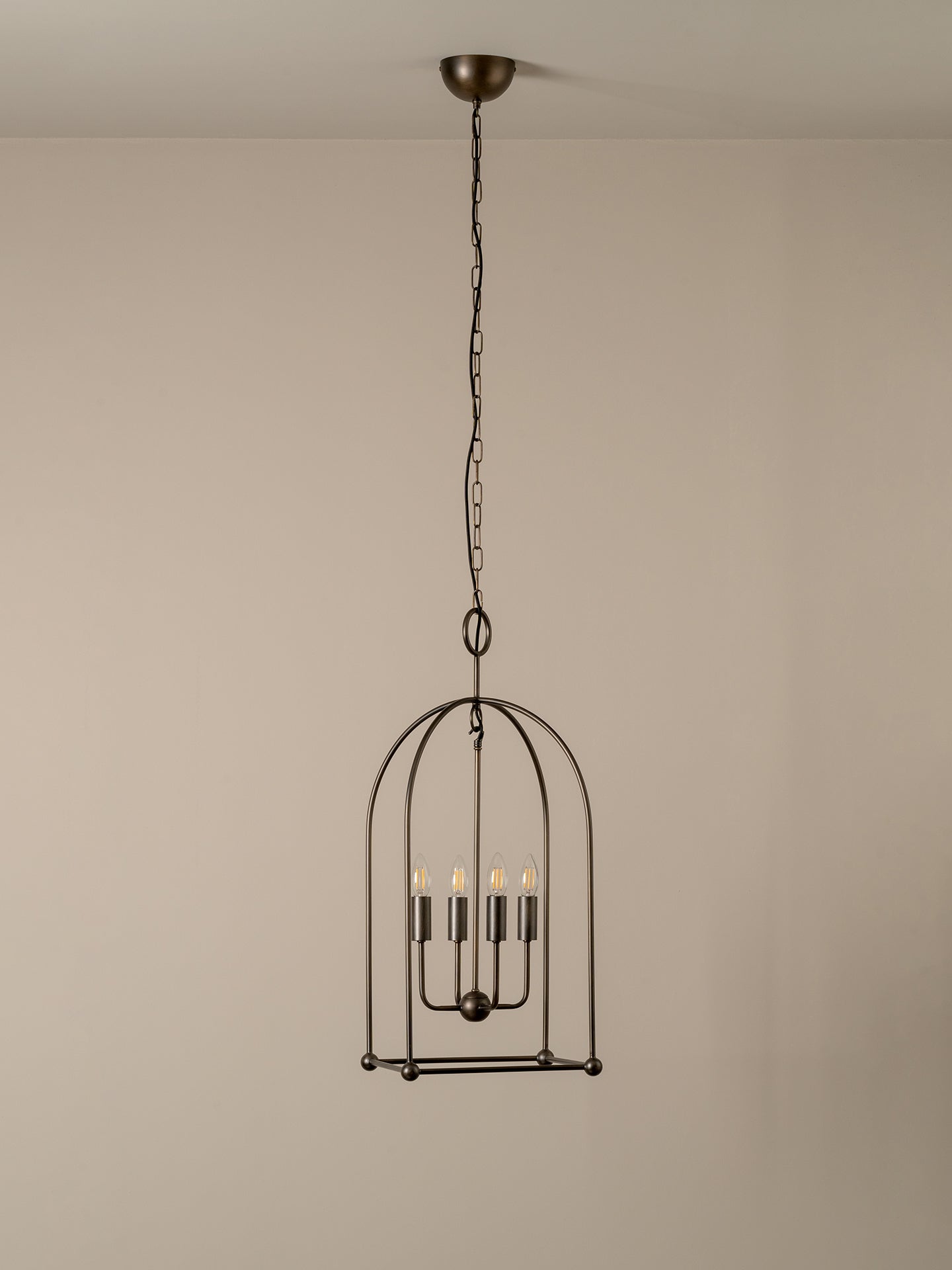 A dark metal birdcage pendant chandelier with five candle-style bulbs, suspended from a ceiling by a chain against a plain beige wall.