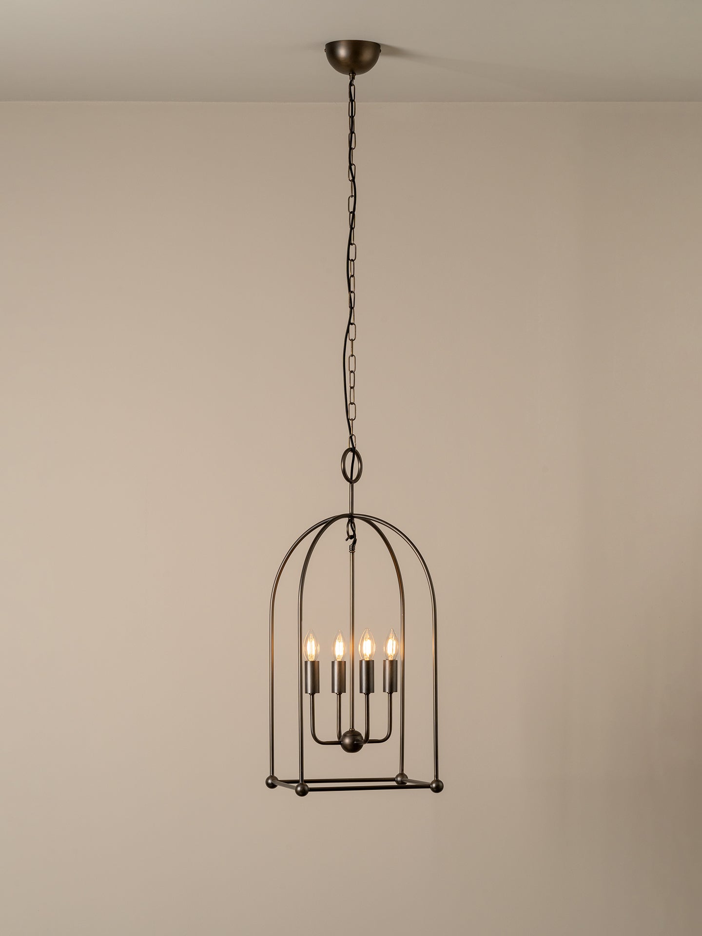 A dark metal birdcage pendant chandelier with five candle-style bulbs, suspended from a ceiling by a chain against a plain beige wall.
