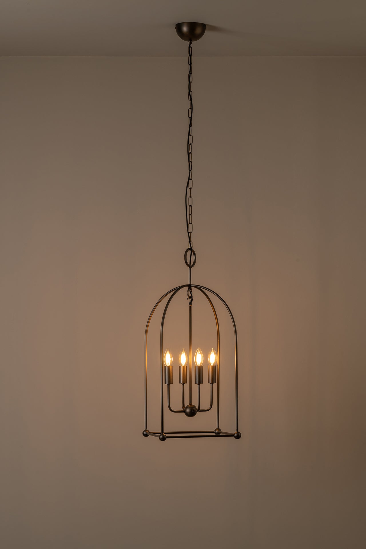 A hanging cage-style pendant light fixture with a dark metal frame and multiple warm glowing bulbs, suspended from a ceiling against a plain beige wall.