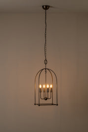 A hanging cage-style pendant light fixture with a dark metal frame and multiple warm glowing bulbs, suspended from a ceiling against a plain beige wall.