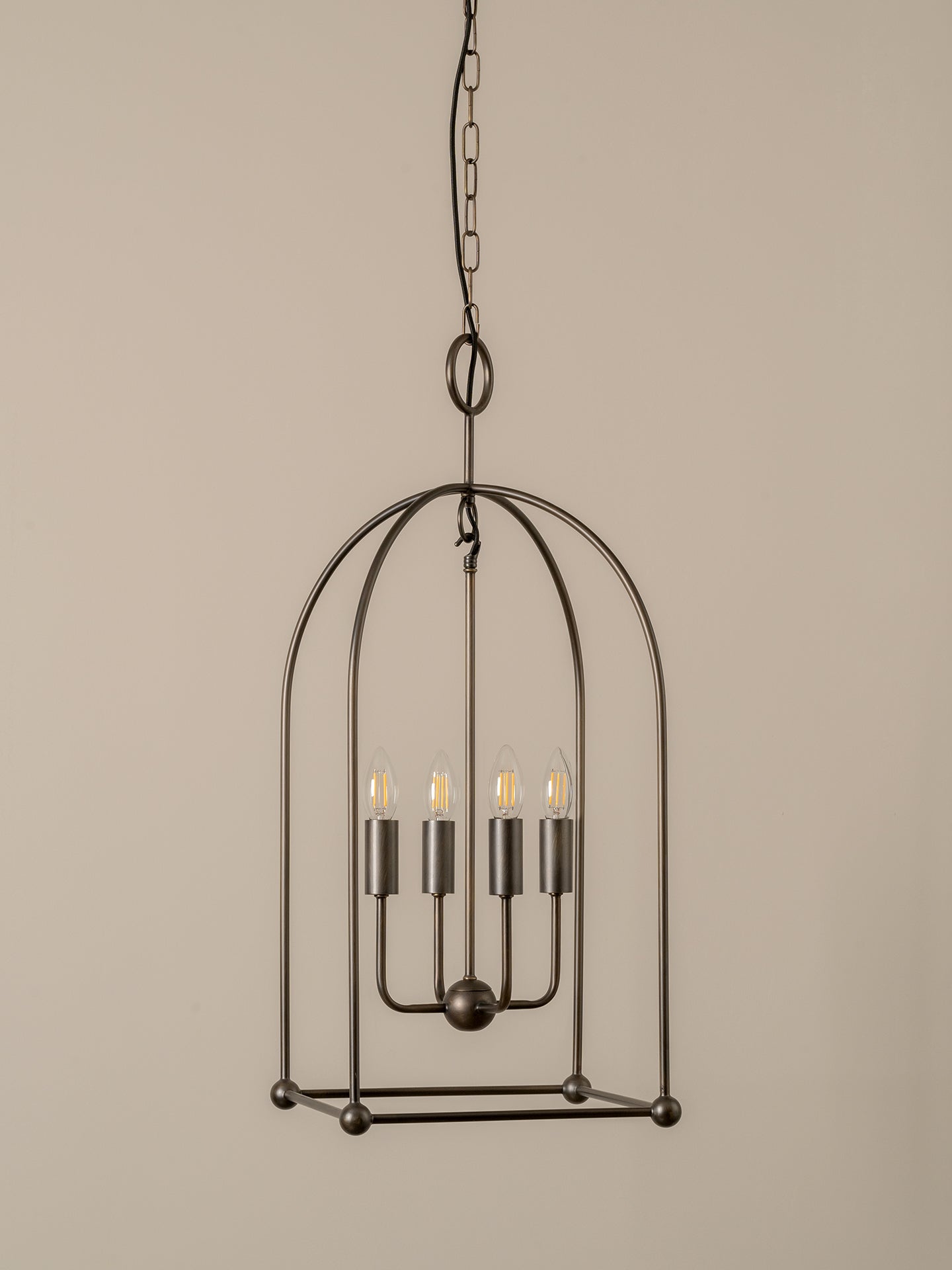 A dark metal cage pendant light fixture with four exposed filament bulbs, suspended by a chain against a plain beige background.