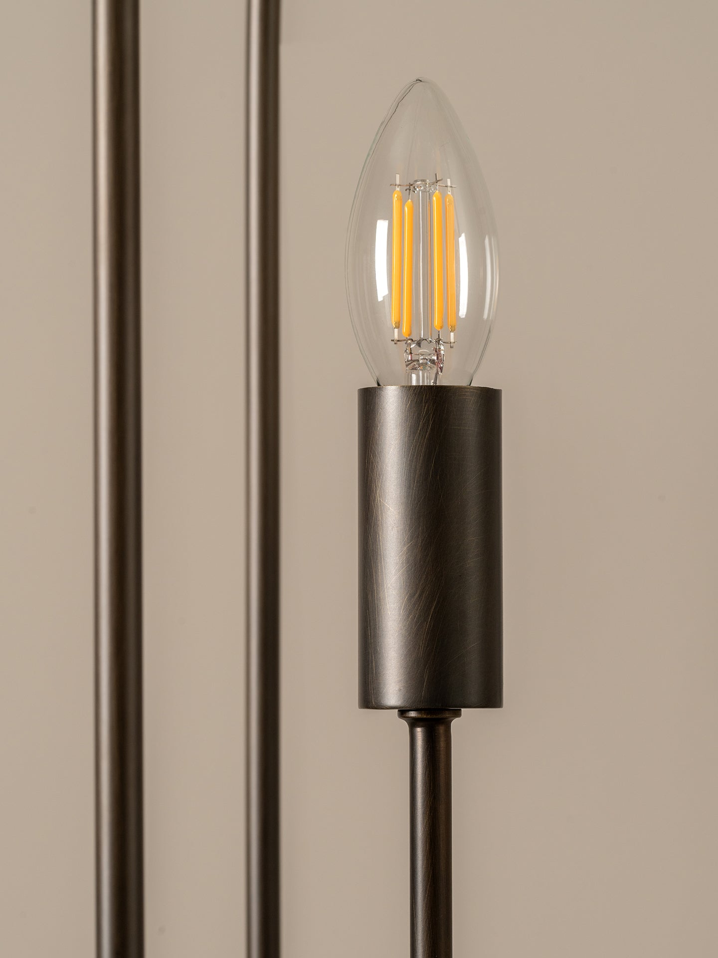 A close-up of a modern wall sconce featuring a brushed dark metal cylindrical shade and a clear glass candle-style bulb with an illuminated warm filament, set against a neutral beige background.