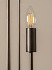 A close-up of a modern wall sconce featuring a brushed dark metal cylindrical shade and a clear glass candle-style bulb with an illuminated warm filament, set against a neutral beige background.
