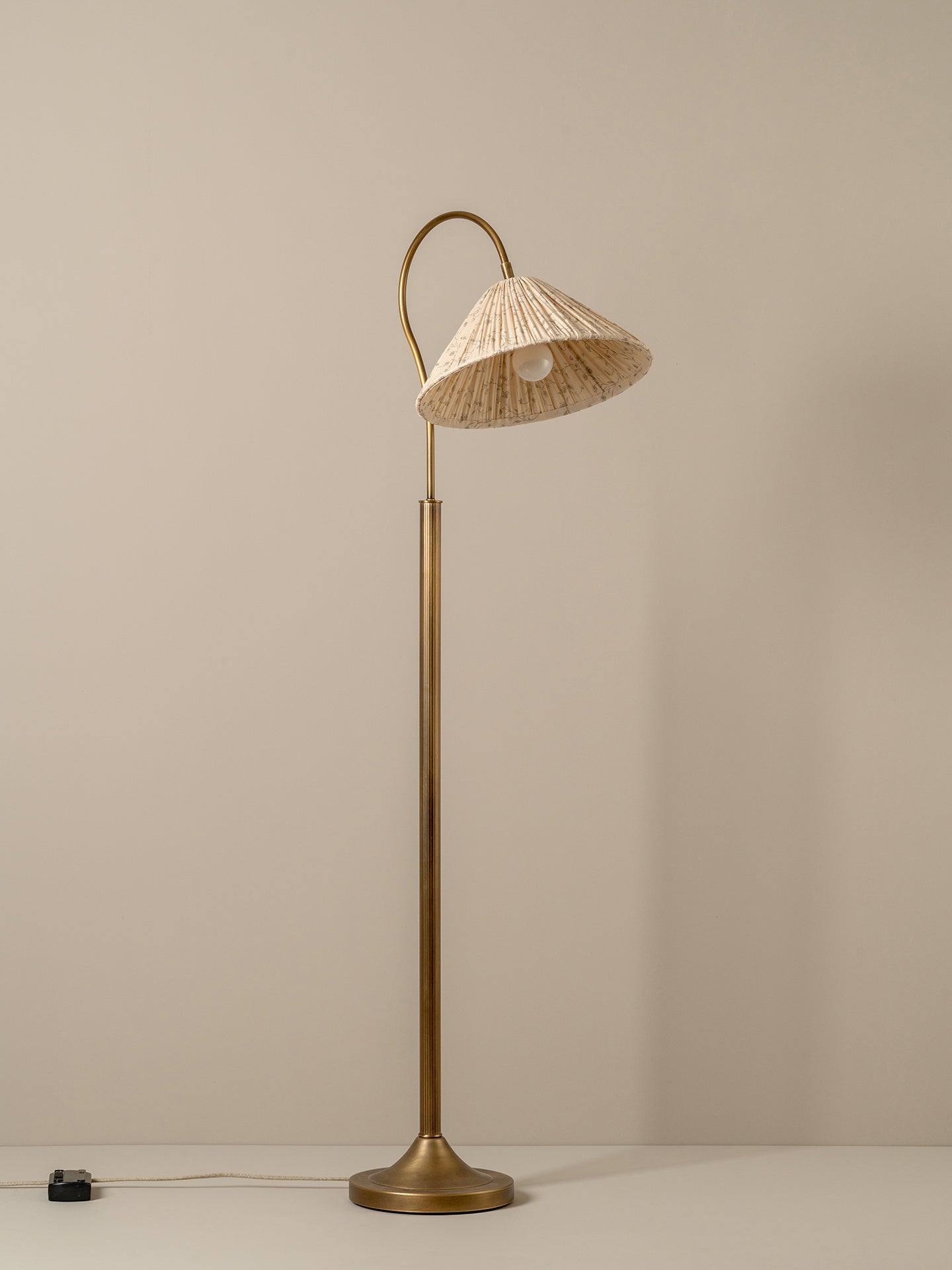A mid-century modern floor lamp with a polished brass base and curved neck, featuring a conical woven straw shade, set against a plain beige wall.