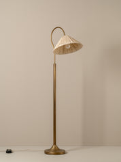 A mid-century modern floor lamp with a polished brass base and curved neck, featuring a conical woven straw shade, set against a plain beige wall.