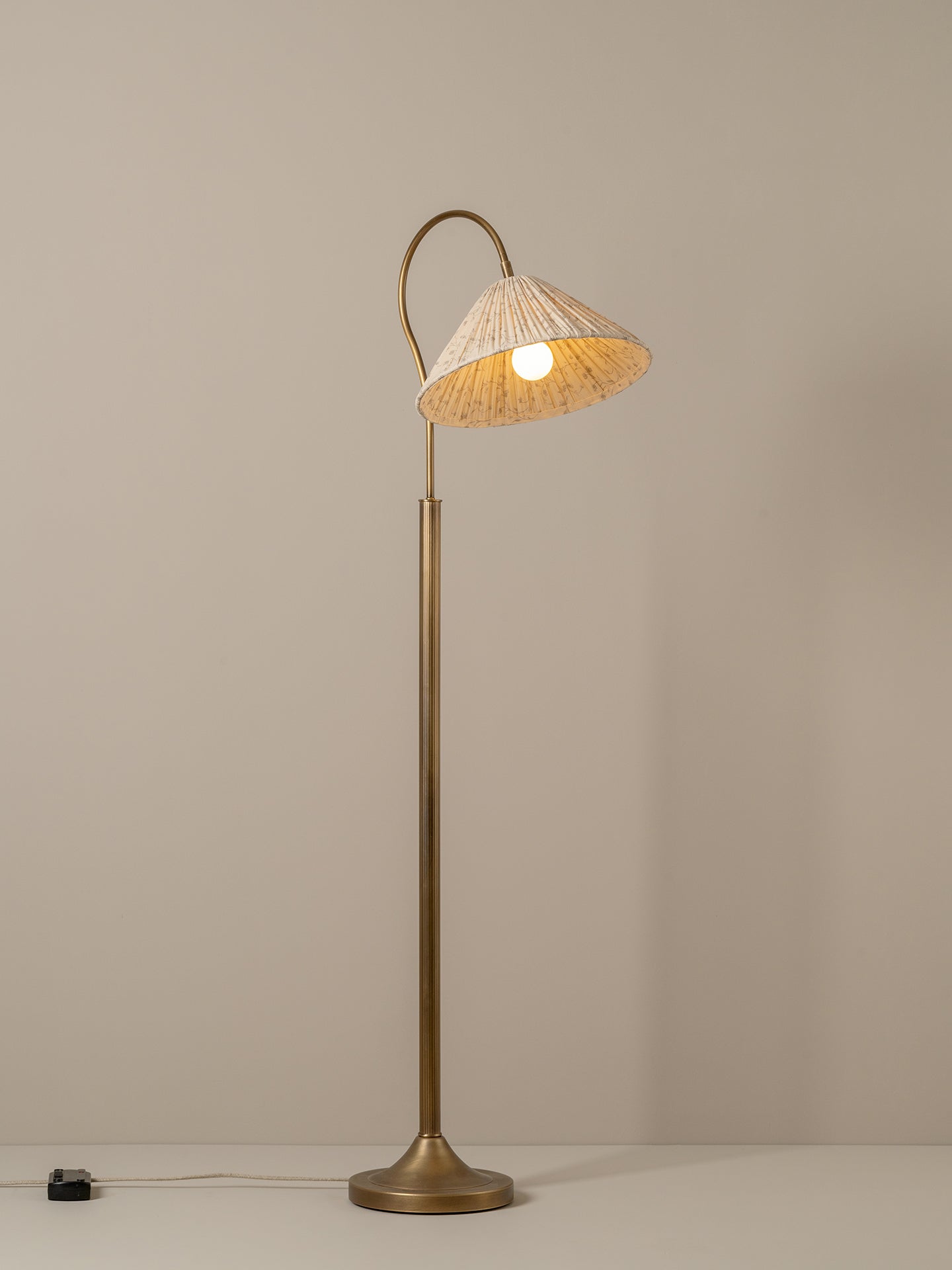 A floor lamp with a tall brass-colored metal stand and curved neck, featuring a conical woven beige shade illuminated from within, set against a plain beige wall.