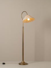 A floor lamp with a tall brass-colored metal stand and curved neck, featuring a conical woven beige shade illuminated from within, set against a plain beige wall.