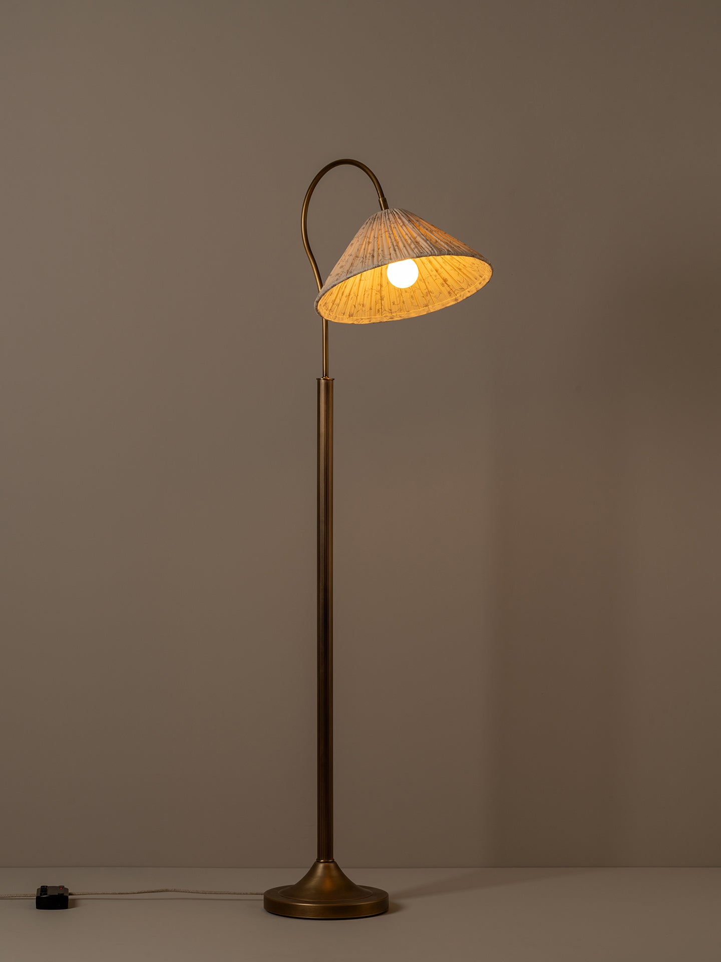 A vintage-style floor lamp with a warm brass finish and a conical shade made of woven natural fiber, glowing with a warm light against a neutral taupe background.