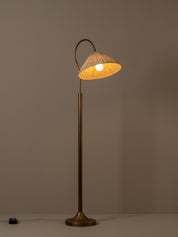 A vintage-style floor lamp with a warm brass finish and a conical shade made of woven natural fiber, glowing with a warm light against a neutral taupe background.