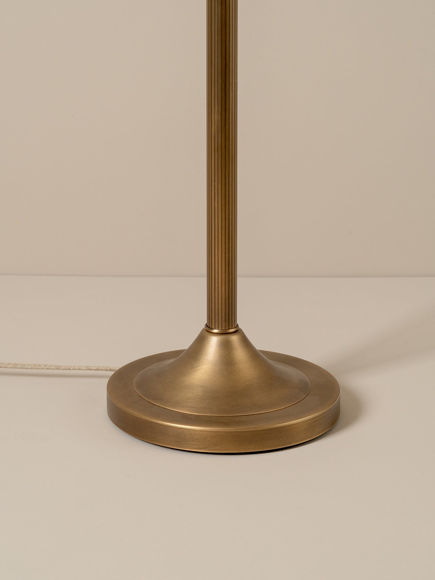 Close-up of a table lamp base and central stem in a brushed brass or antique gold finish, photographed against a plain warm beige background.