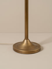Close-up of a table lamp base and central stem in a brushed brass or antique gold finish, photographed against a plain warm beige background.