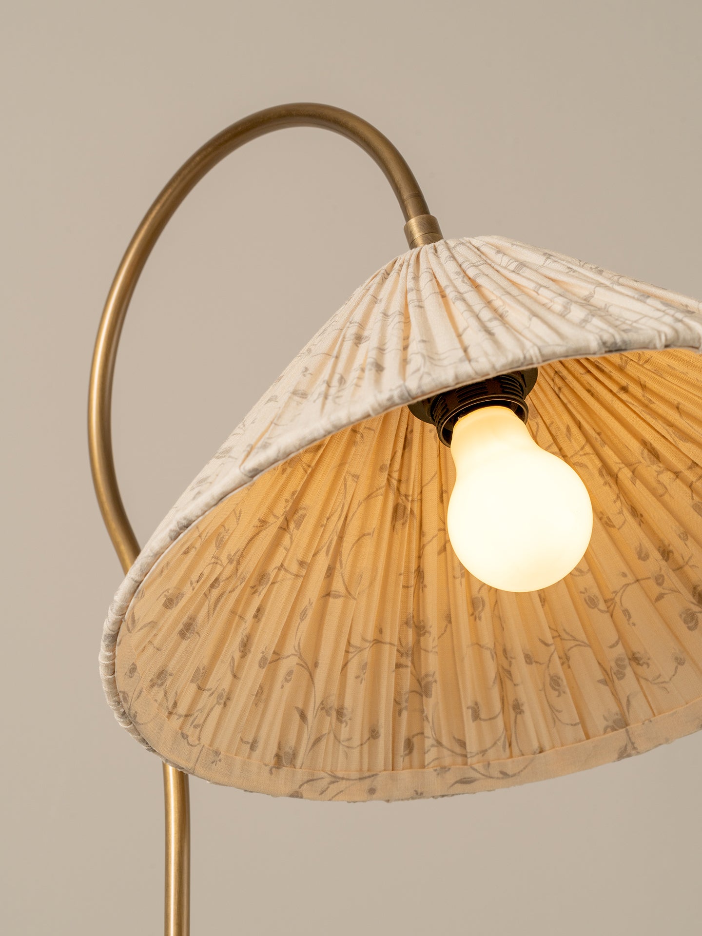 A close-up of a floor lamp with a curved brass metal stand and a pleated fabric shade in cream and beige with a subtle floral pattern, illuminated by a warm light from a visible bulb.