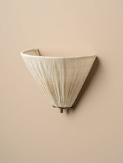 A modern wall sconce with a pleated, cone-shaped shade in a warm off-white or cream tone, mounted on a solid beige wall.
