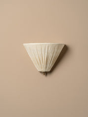 A modern wall sconce with a pleated, cone-shaped shade in a warm off-white or cream color, mounted on a solid beige wall.
