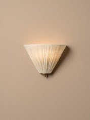 A modern wall sconce with a pleated, conical shade made of a warm off-white fabric, shown illuminated against a solid beige wall.