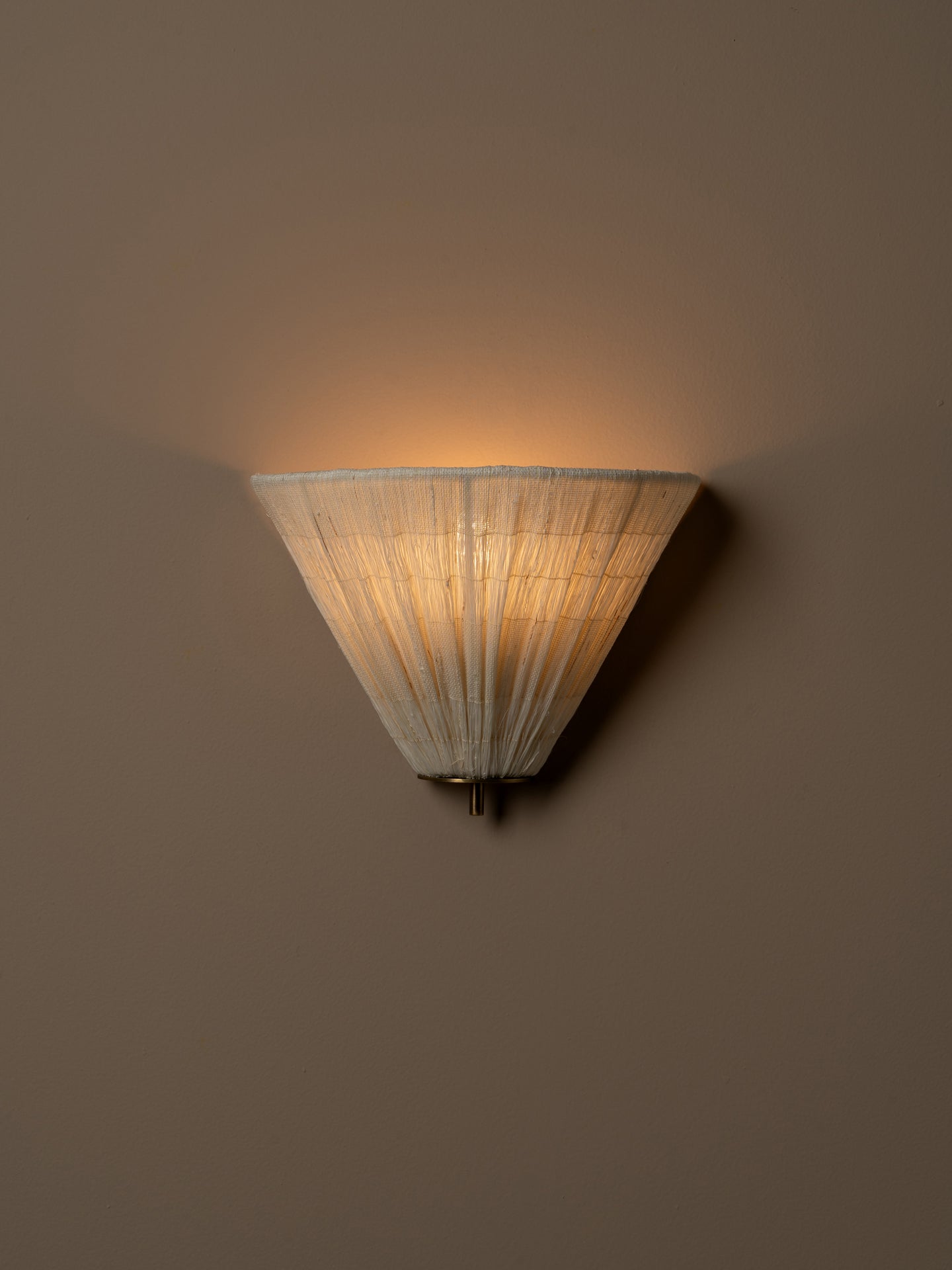 A modern wall sconce with a pleated, conical shade made of a warm off-white fabric, shown illuminated against a solid beige wall. light turned on.