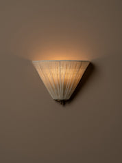 A modern wall sconce with a pleated, conical shade made of a warm off-white fabric, shown illuminated against a solid beige wall. light turned on.