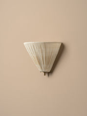 A modern wall sconce with a pleated, cone-shaped shade in a warm off-white or cream tone, mounted on a solid beige wall.
