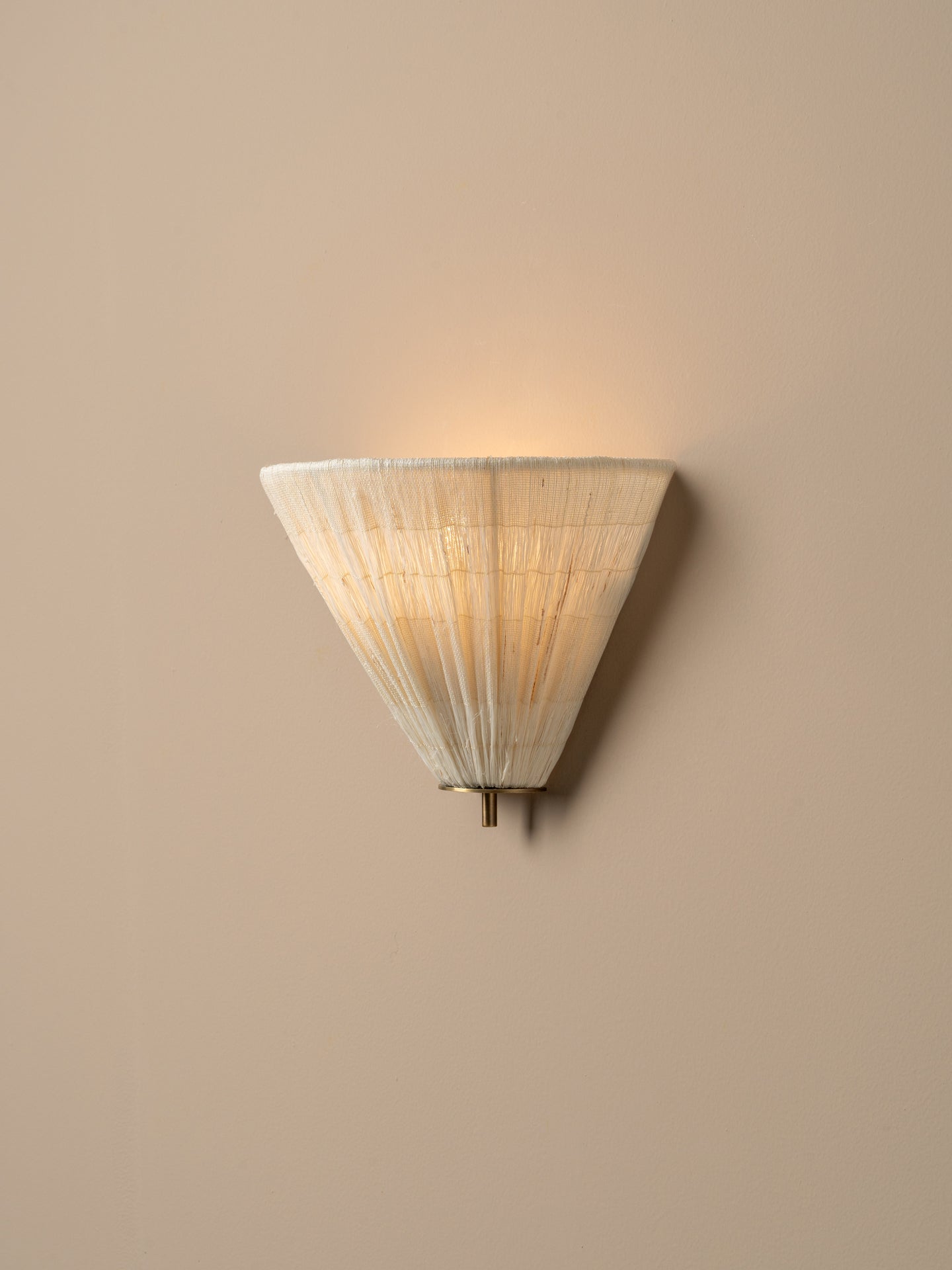 A modern wall sconce with a conical, pleated shade made of a warm off-white material, glowing with a soft warm light against a solid beige wall.