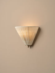 A modern wall sconce with a conical, pleated shade made of a warm off-white material, glowing with a soft warm light against a solid beige wall.