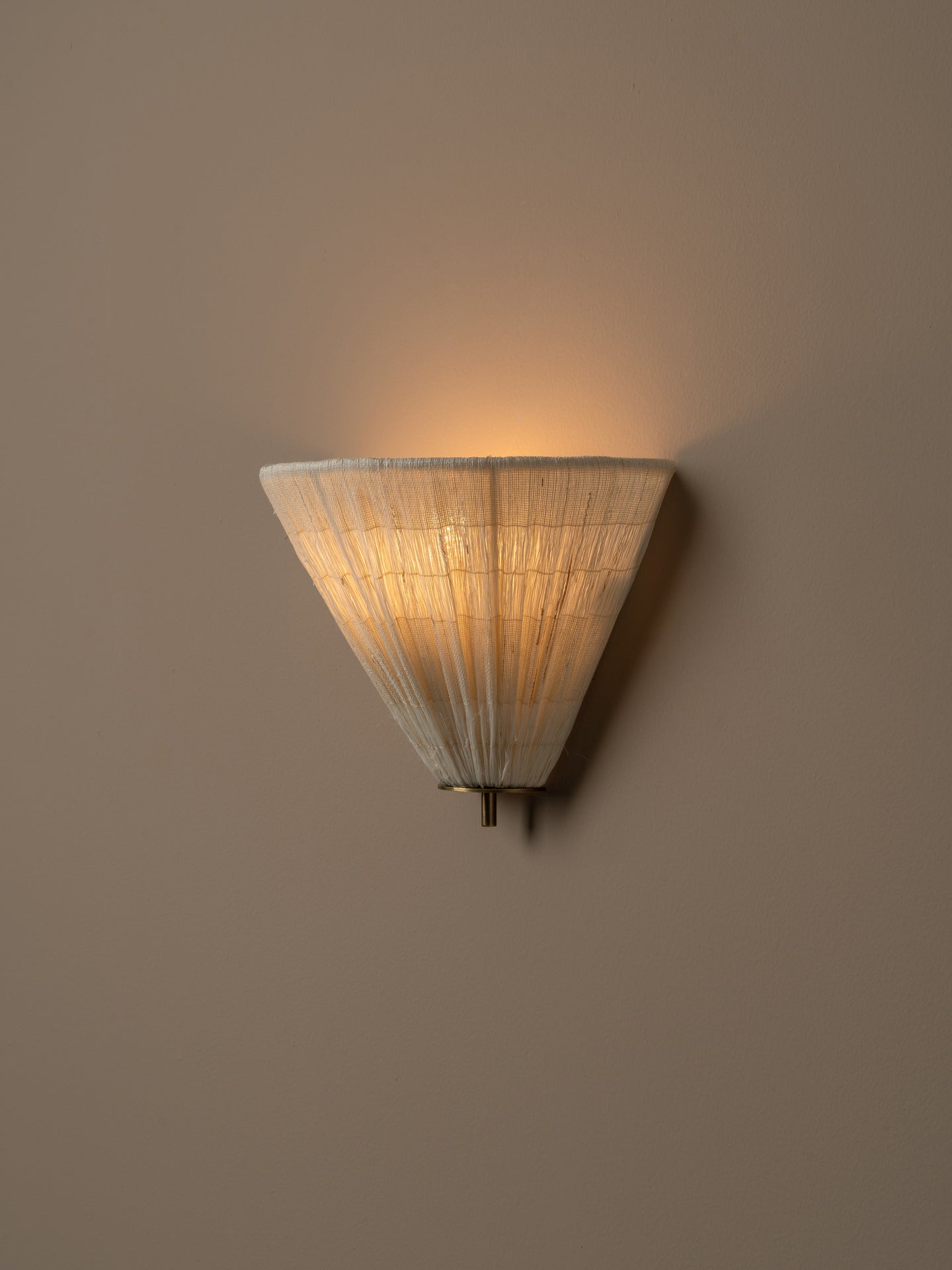 A modern wall sconce with a tapered, pleated shade made of a light natural fiber material, glowing with a warm yellow light against a solid taupe wall.