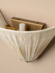 Close-up of a wall-mounted sconce with a pleated cream fabric shade and a brushed brass metal backplate against a warm beige wall.