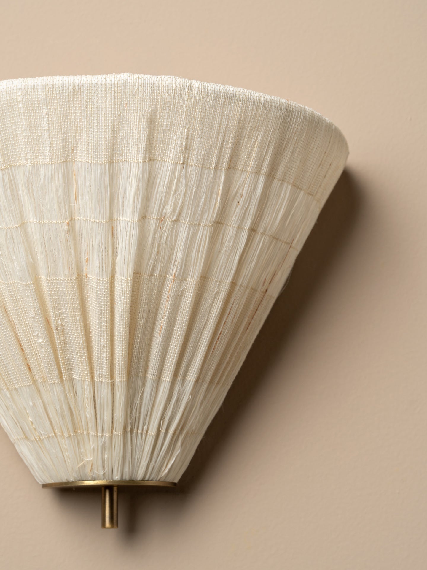 A close-up of a wall-mounted sconce with a pleated, textured off-white shade and a small brass-toned metal finial against a warm beige wall.