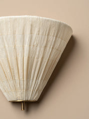 A close-up of a wall-mounted sconce with a pleated, textured off-white shade and a small brass-toned metal finial against a warm beige wall.