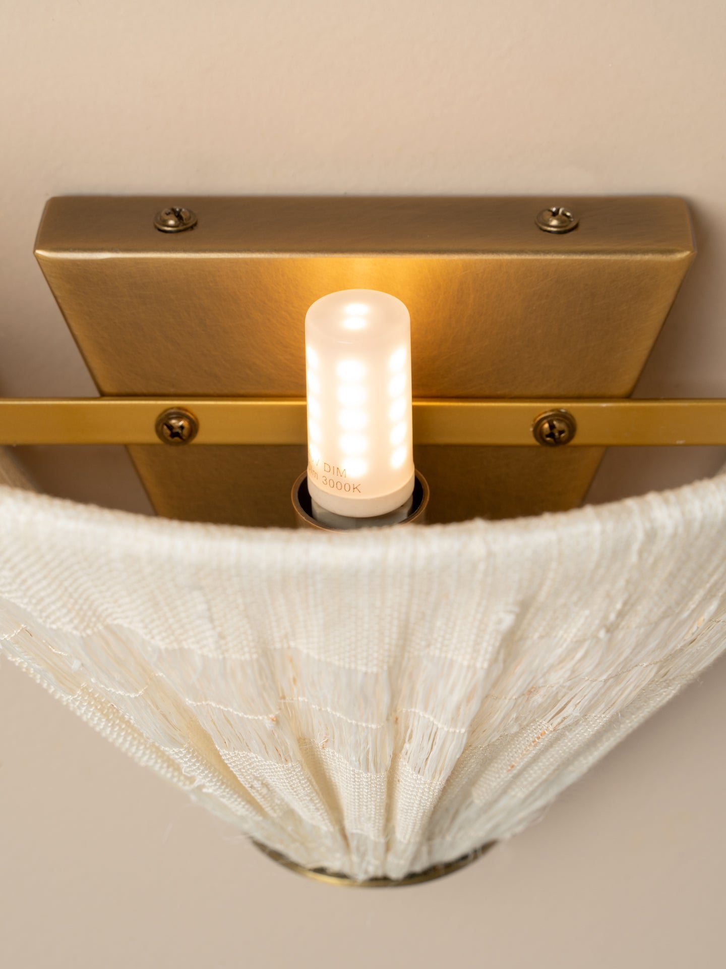 A close-up of a wall-mounted light fixture with a brushed brass backplate, a horizontal brass bar, a glowing cylindrical bulb, and a white textured fabric shade draping downward against a beige wall.