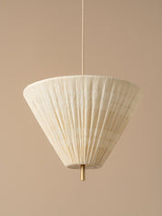 A modern pendant light with a pleated, cone-shaped off-white shade suspended from a brass ceiling canopy against a warm beige wall.