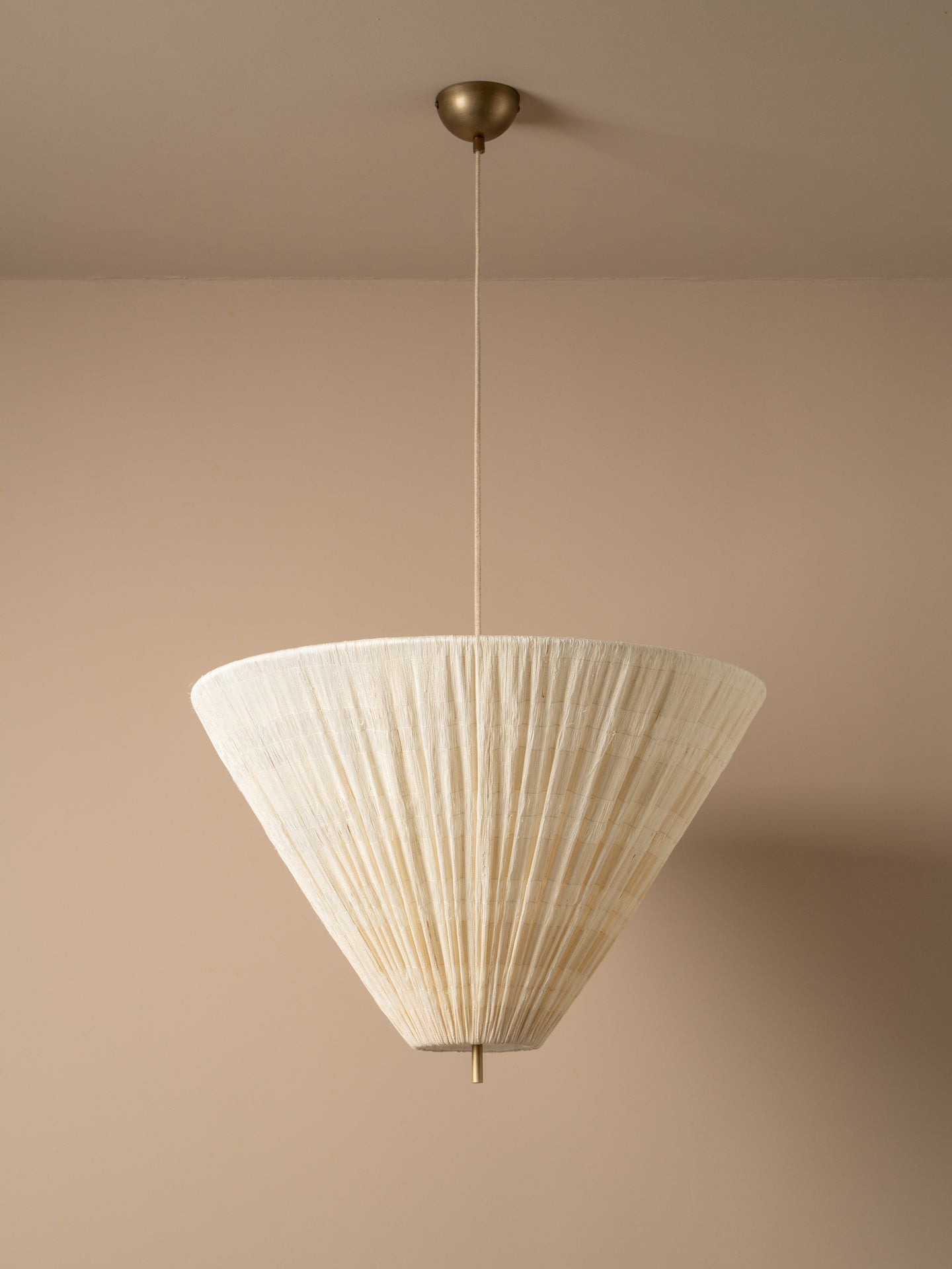 A modern pendant light with a pleated, cone-shaped off-white shade suspended from a brass ceiling canopy against a warm beige wall.
