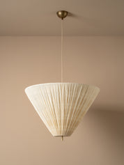 A modern pendant light with a pleated, cone-shaped off-white shade suspended from a brass ceiling canopy against a warm beige wall.