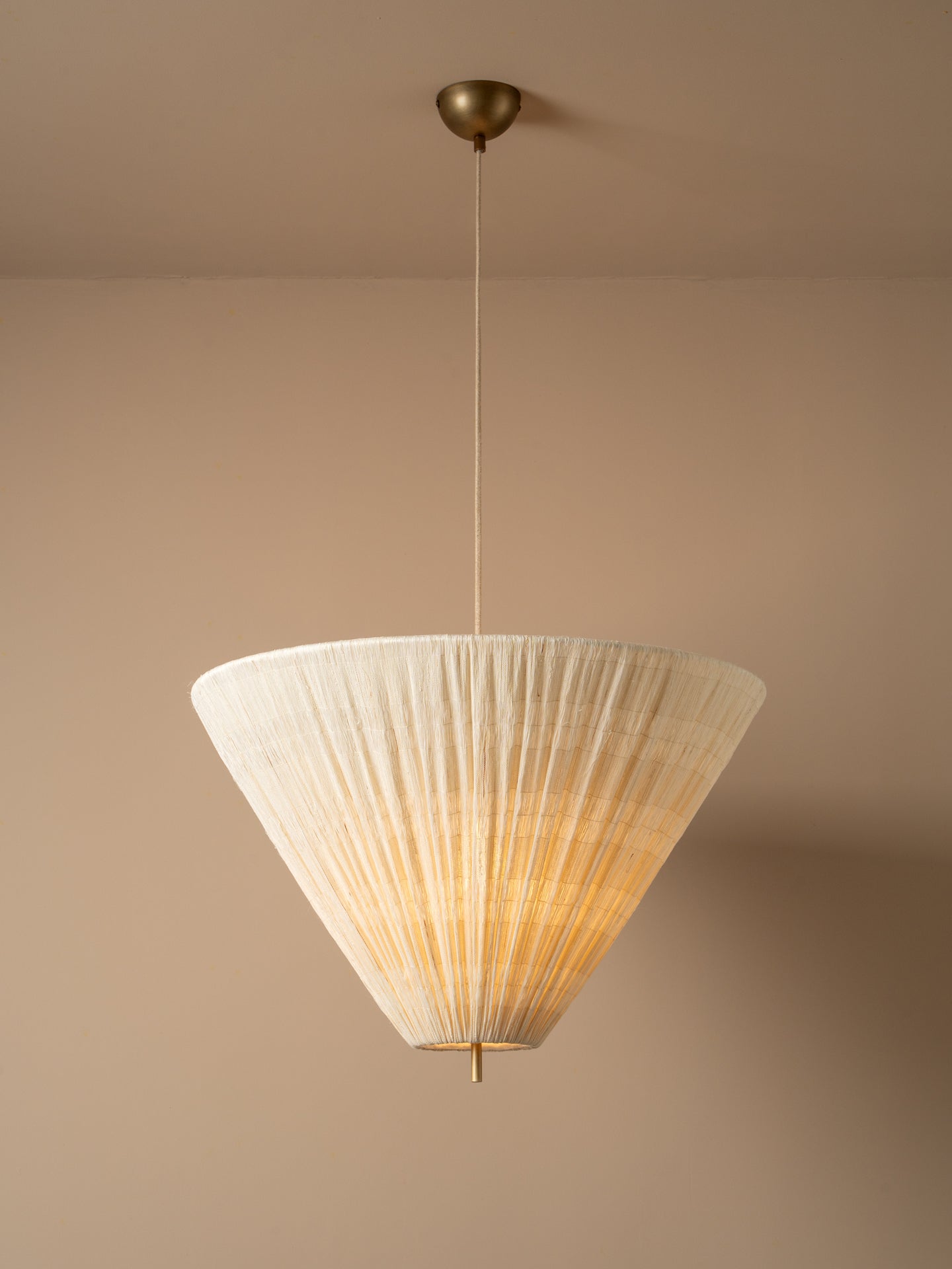A modern pendant light with a pleated, cone-shaped off-white shade suspended from a brass ceiling canopy against a warm beige wall.