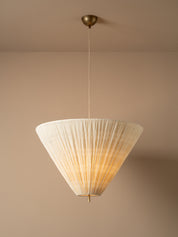 A modern pendant light with a pleated, cone-shaped off-white shade suspended from a brass ceiling canopy against a warm beige wall.