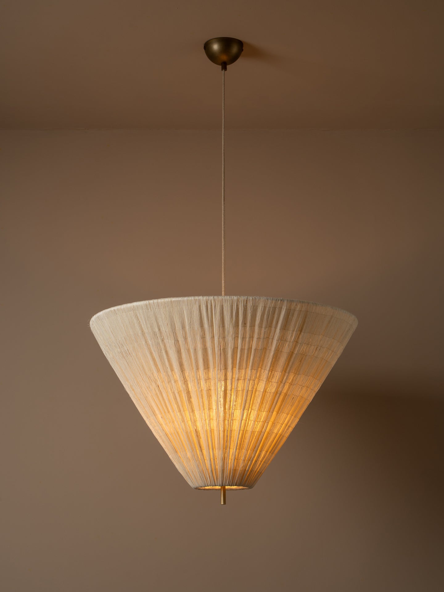 A modern pendant light with a pleated, cone-shaped off-white shade suspended from a brass ceiling canopy against a warm beige wall.