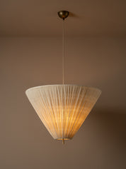 A modern pendant light with a pleated, cone-shaped off-white shade suspended from a brass ceiling canopy against a warm beige wall.