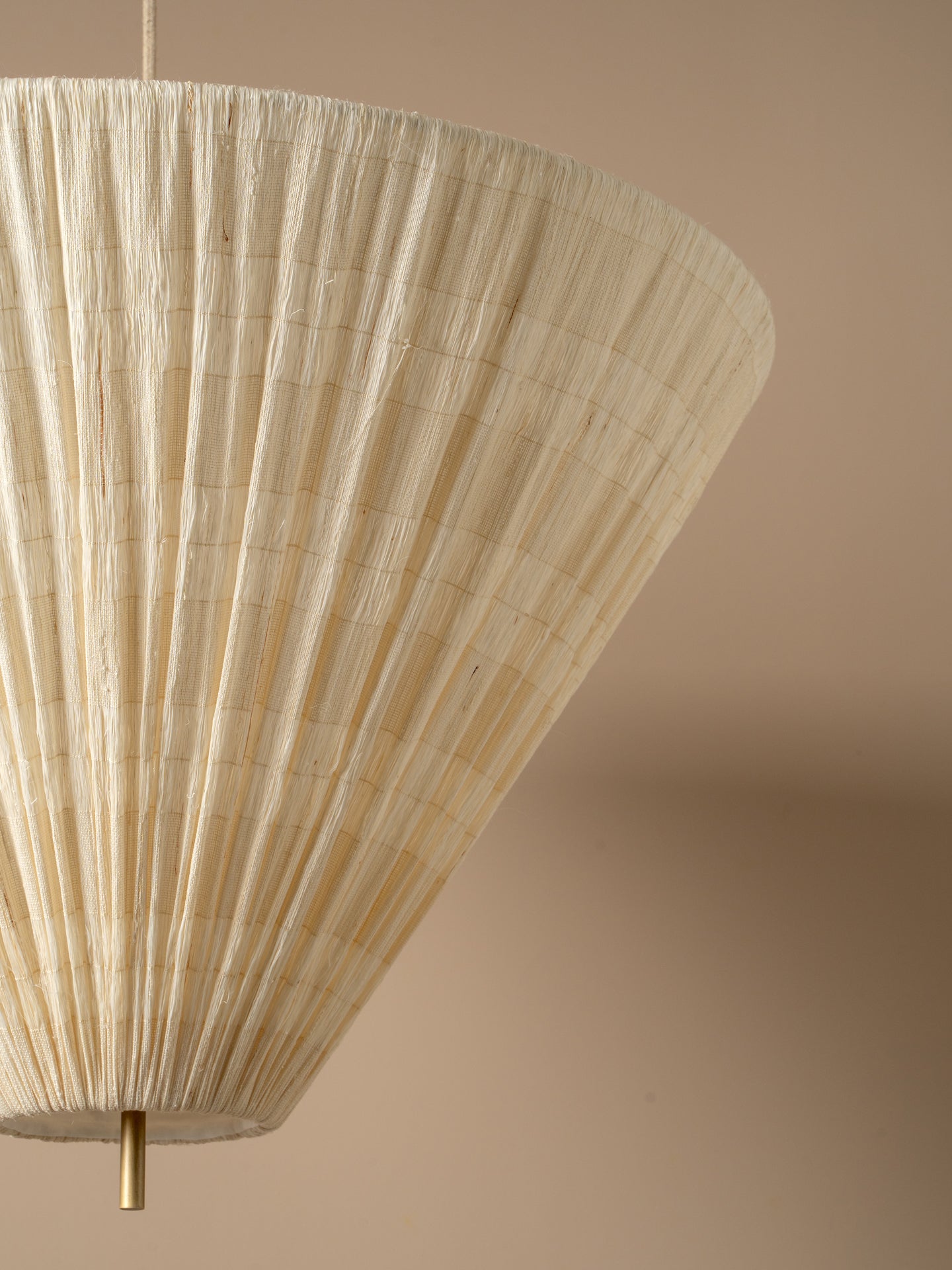A close-up of a pleated conical pendant lampshade upside down in a warm cream or off-white tone, suspended by slim brass-toned metal rods against a solid beige background.