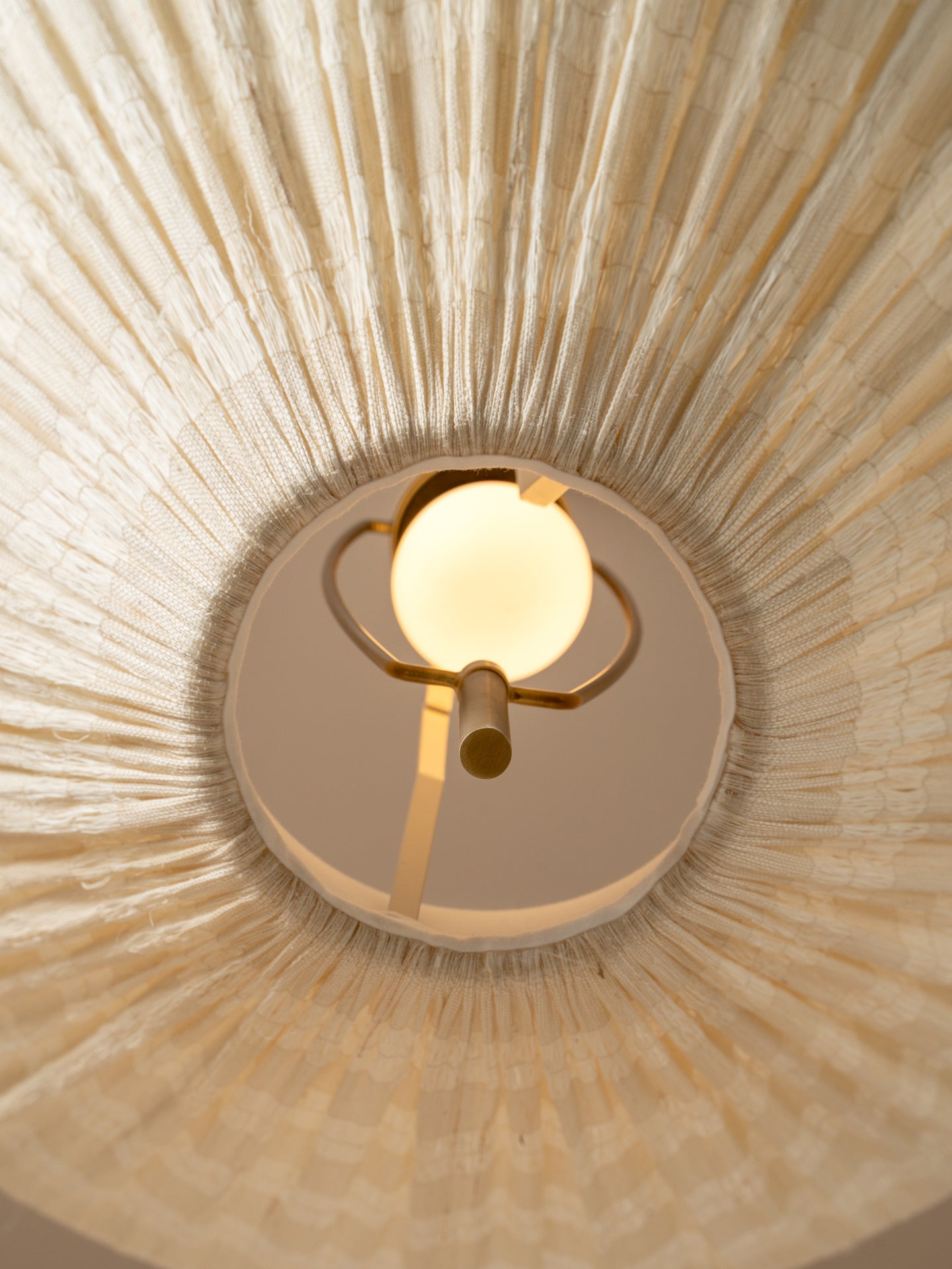 A close-up, upward view of a ceiling light fixture installed in a circular opening, showing a warm glowing globe bulb and brass-toned metal arms set against a textured beige radial ceiling surface.