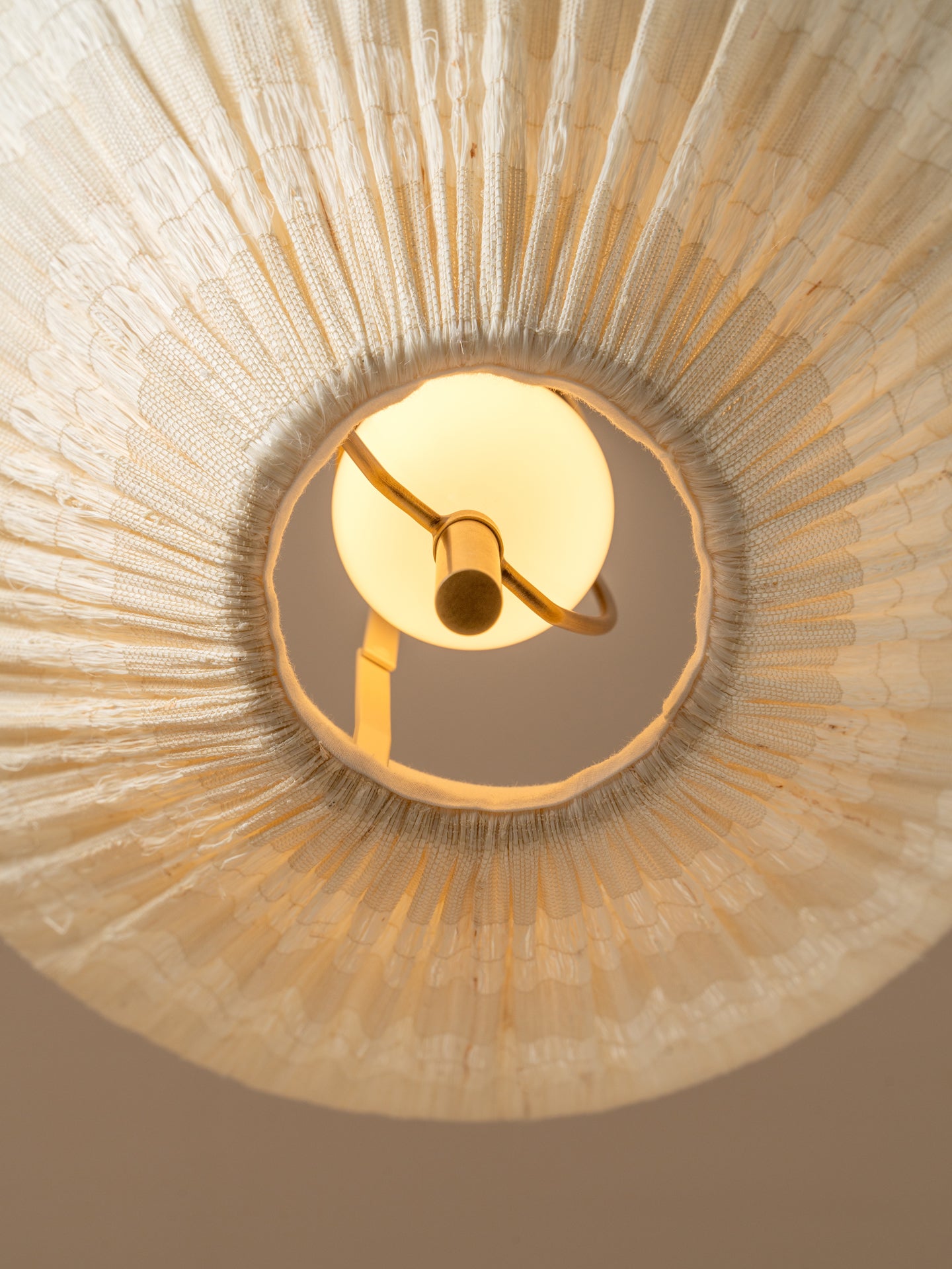 A close-up, upward view of a ceiling light fixture installed in a circular opening, showing a warm glowing globe bulb and brass-toned metal arms set against a textured beige radial ceiling surface.