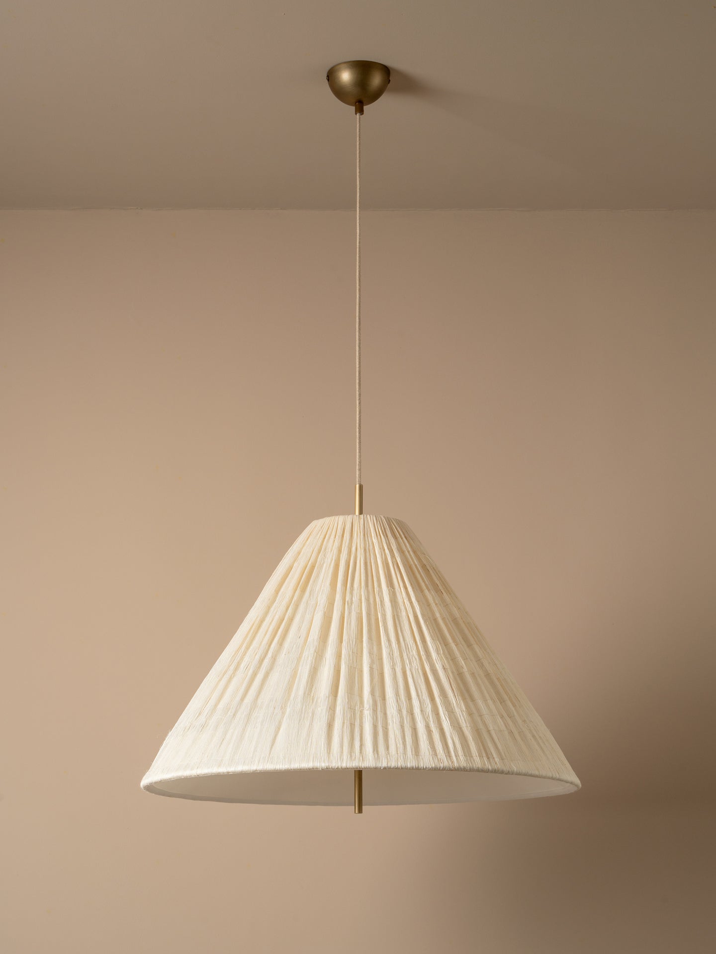A modern pendant light with a pleated, cone-shaped off-white shade suspended from a brass ceiling canopy against a warm beige wall.