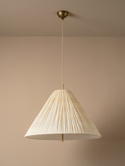 A modern pendant light with a pleated, cone-shaped off-white shade suspended from a brass ceiling canopy against a warm beige wall.