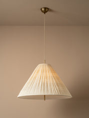 A modern pendant light with a pleated, cone-shaped off-white shade suspended from a brass ceiling canopy against a warm beige wall.