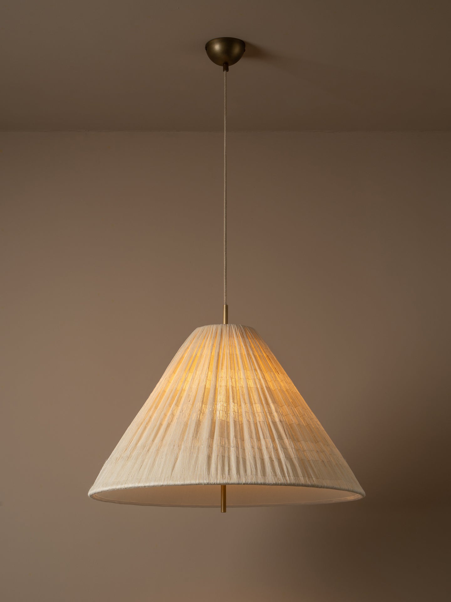 A modern pendant light with a pleated, cone-shaped off-white shade suspended from a brass ceiling canopy against a warm beige wall.