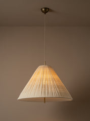 A modern pendant light with a pleated, cone-shaped off-white shade suspended from a brass ceiling canopy against a warm beige wall.