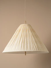 A modern pendant light with a pleated, cone-shaped off-white shade suspended from a brass ceiling canopy against a warm beige wall.